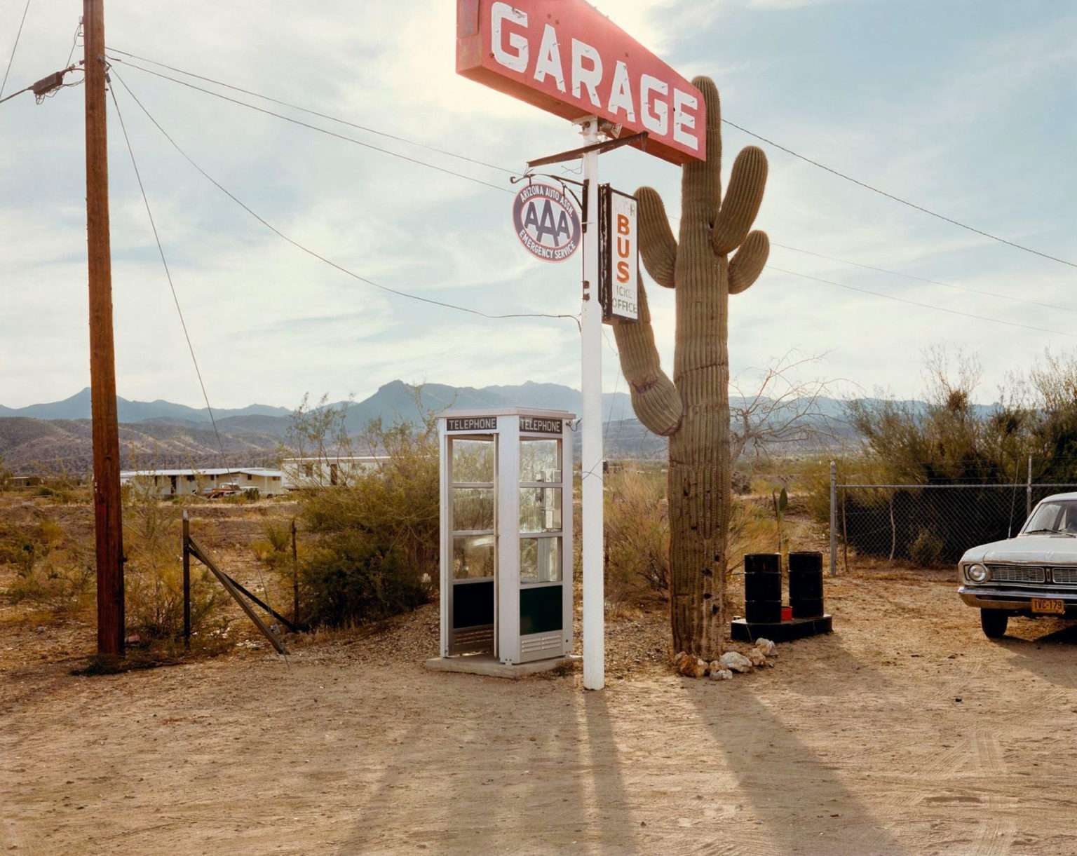 Photo d'un paysage désertique avec une cabine téléphonique, un grand cactus et un panneau "Garage". Une vieille voiture est garée à côté.
