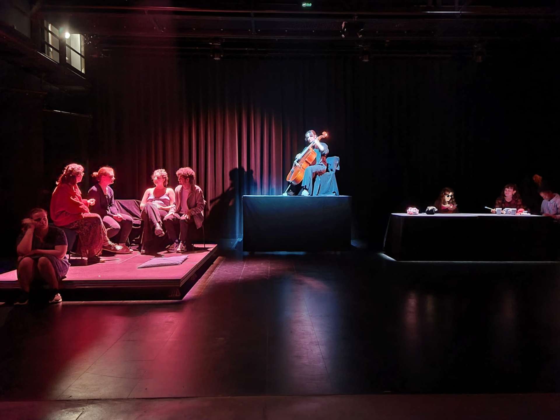 Photo d'une pièce de théâtre. Deux groupes de personnes sont assis de chaque côtés de la scène. Au centre, une femme est assise sur une estrade avec un violoncelle. 