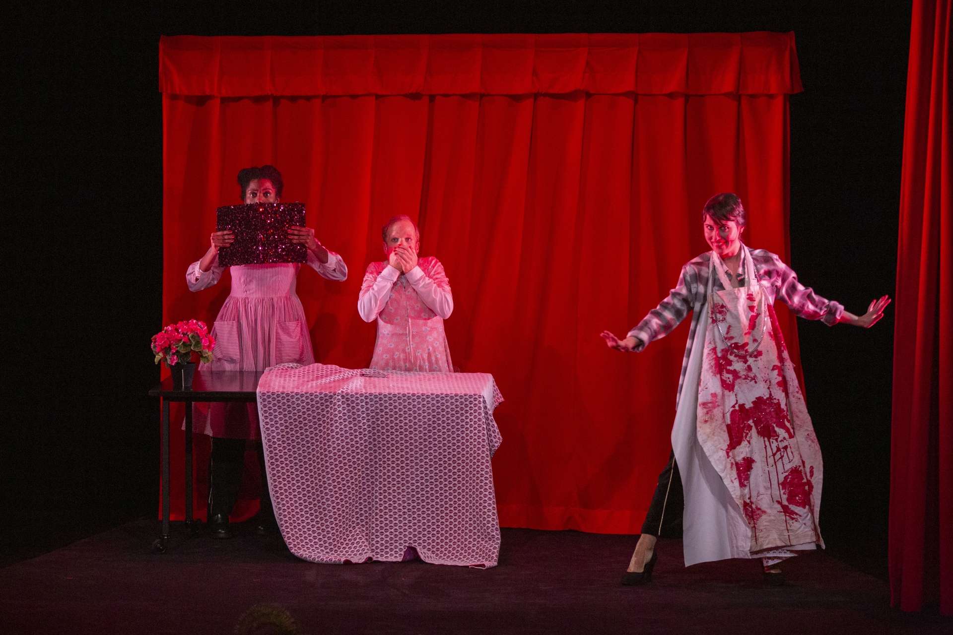 Photo du spectacle Fin (Faim) : Trois personnes sont sur scènes, deux d'entre elles sont derrière une table et se cache le visage. Elles semblent avoir peur. La troisième personne à droite de l'image à un tablier taché de sang.