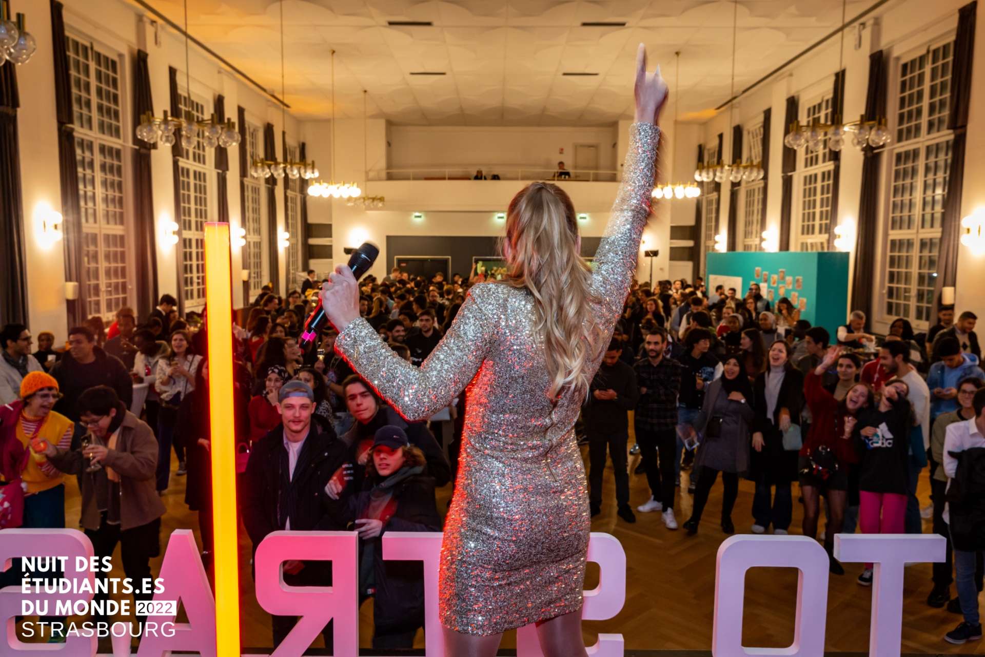 Photo de la Nuit des étudiant·e·s du monde. Une jeune femme en robe de soirée à paillettes, un micro à la main et l'autre main levée en l'air, tourne le dos au photographe et fait face à une foule d'étudiant·e·s internationaux.