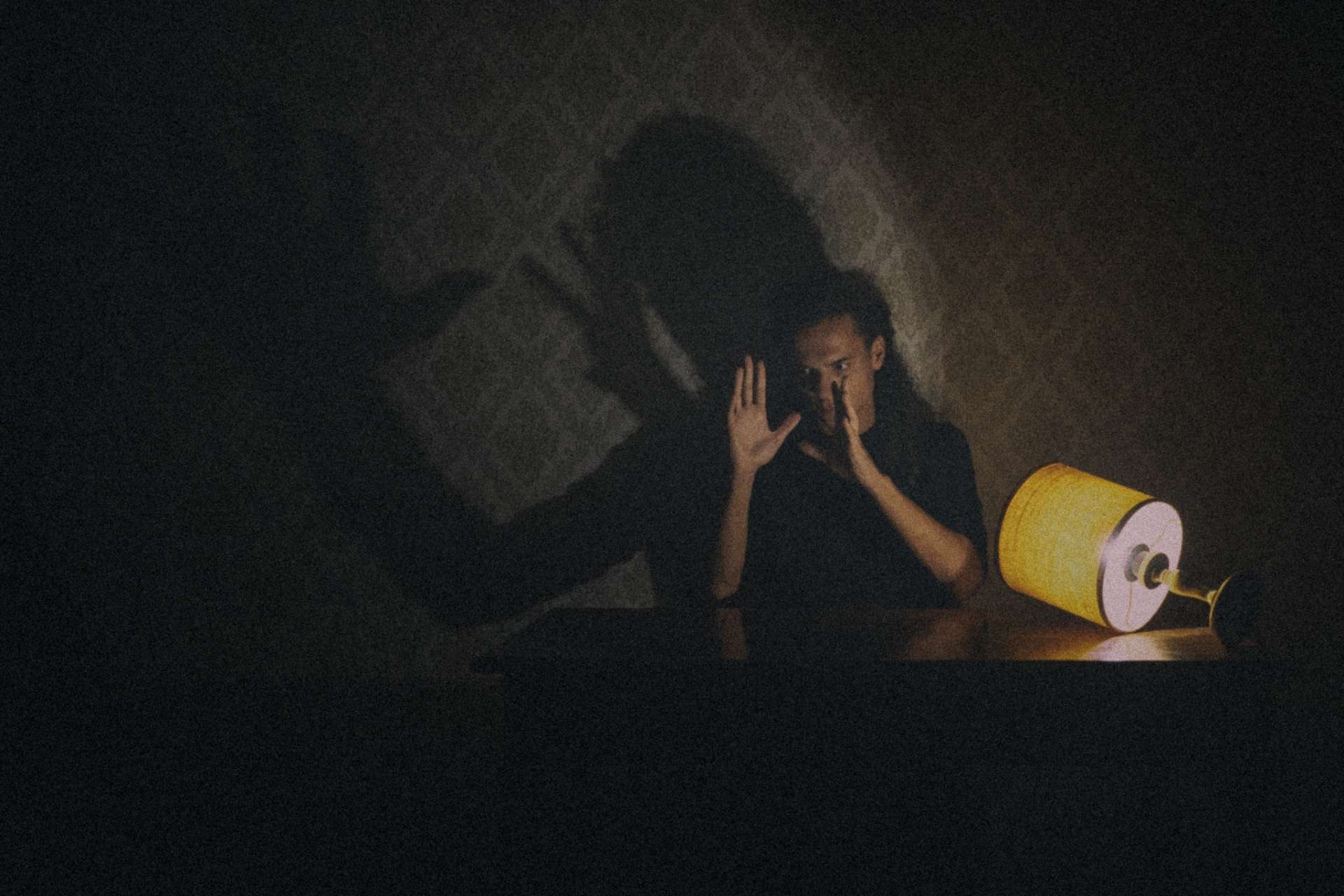 L'acteur est assis derrière une table et, à la lueur d'une petite lampe à abat-jour jaune couchée, forme une ombre avec ses mains et sa tête sur la tapisserie derrière lui.